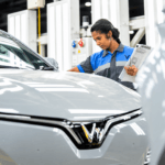 Woman inspecting a silver electric car in an automotive factory while holding a clipboard