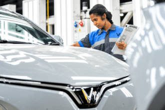 Woman inspecting a silver electric car in an automotive factory while holding a clipboard
