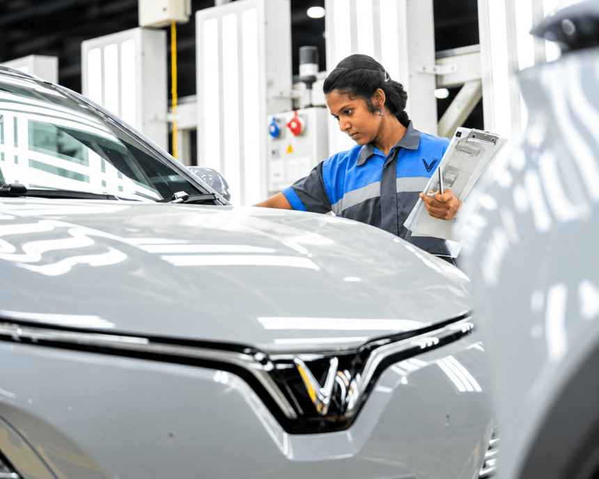 Woman inspecting a silver electric car in an automotive factory while holding a clipboard